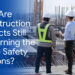 Two construction workers wearing white hard hats stand on an unfinished concrete building level, looking out over a construction site filled with metal rebar. One worker wears a yellow high-visibility vest, while the other holds a digital tablet. Overlaid on a blue background on the left side of the image is large text that reads, "Why Are Construction Projects Still Relearning the Same Safety Lessons?".